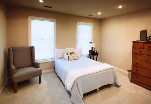 A simple bedroom with a single bed covered in a light gray quilt and white pillows, one pillow featuring a flamingo design. There is a gray upholstered armchair to the left of the bed, two windows with white blinds behind the bed, a small black side table with a lamp and decorative items on the right side, and a wooden dresser with a TV on top on the right side of the room. The walls are beige and the floor is carpeted in a light color.