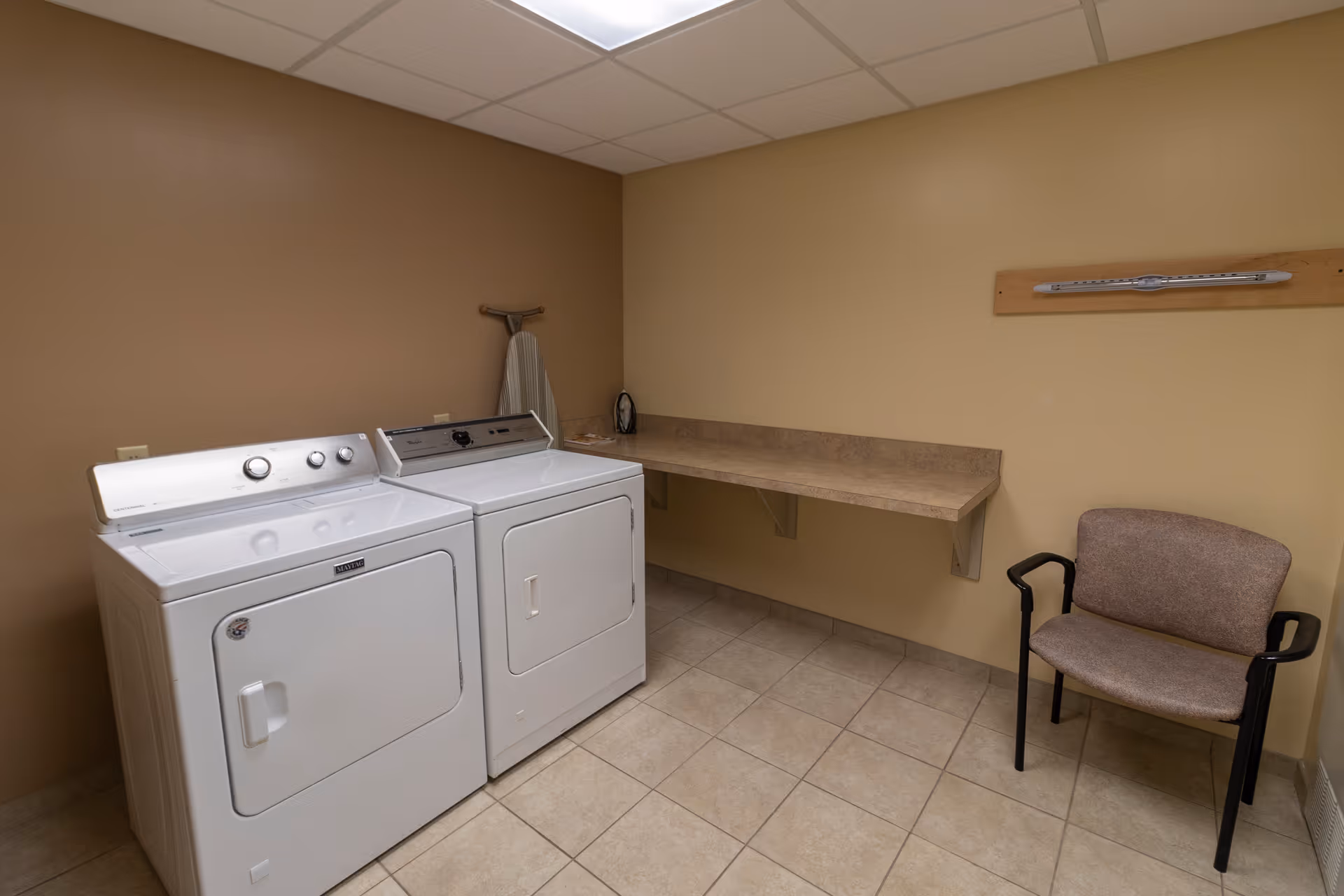 A laundry room with a white washing machine and dryer side by side against a beige wall. There is a countertop above the machines extending along the wall, an ironing board leaning against the wall, an iron on the countertop, and a beige chair with black armrests in the corner. The floor is tiled and the ceiling has fluorescent lighting.