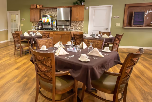 Dining area in an assisted living facility with several tables covered in dark brown tablecloths, each set with white napkins, cups, glasses, and silverware. The room has wooden chairs with patterned cushions, wood flooring, and a beverage station with juice dispensers and a coffee machine against a wall with wooden cabinets and a tiled backsplash.