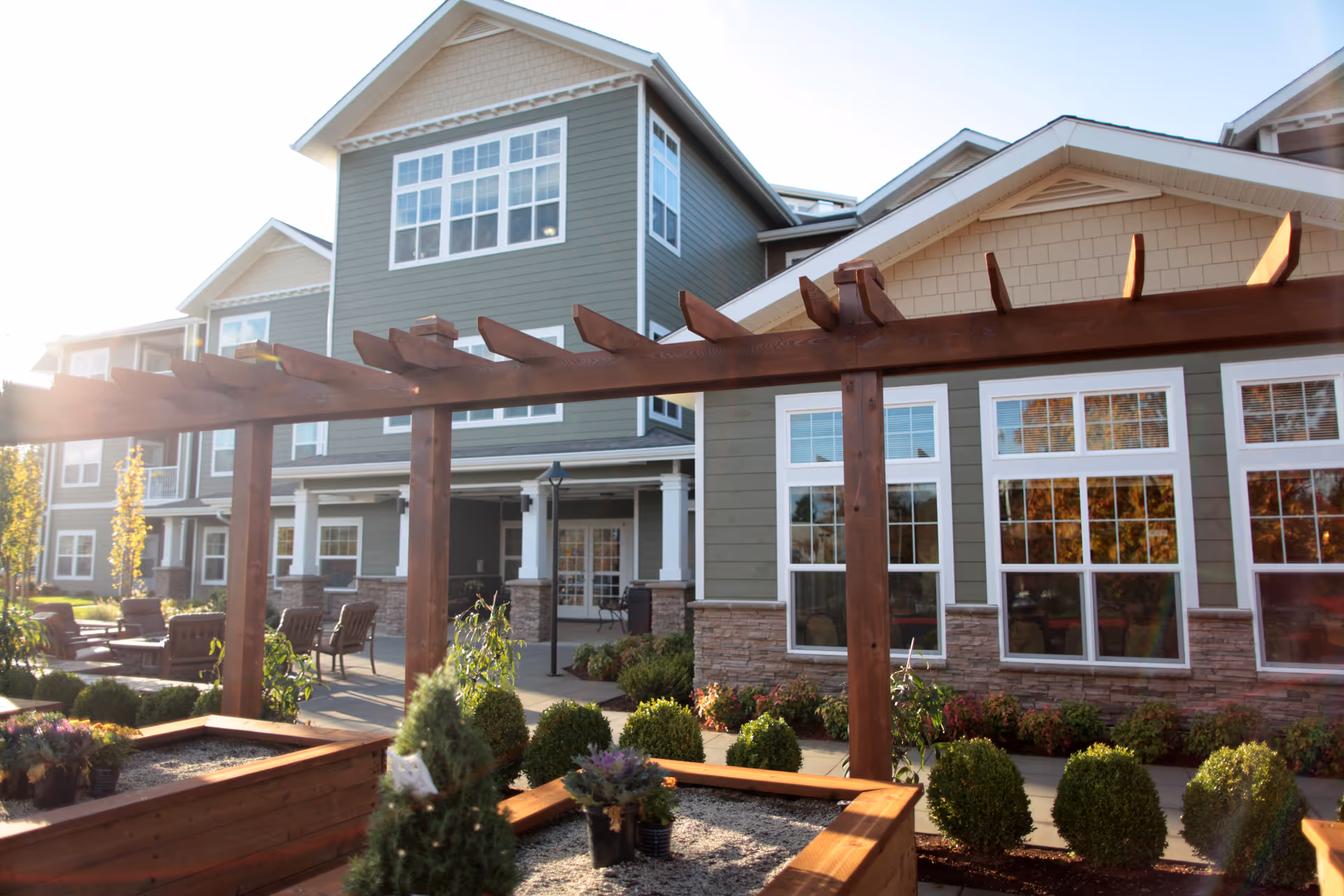 Outdoor patio area of a senior living facility with wooden pergola, raised garden beds, small shrubs, and seating arrangements. The building exterior features large windows, stone accents, and green siding under a clear sky with sunlight.