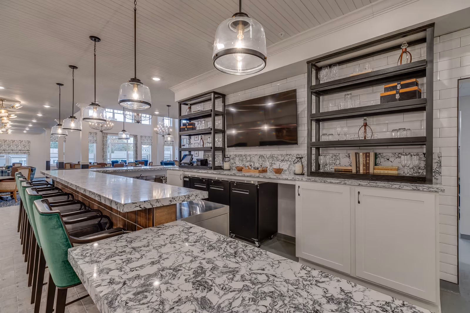 Interior view of a modern dining and bar area in a senior living facility with marble countertops, green bar stools, pendant lights, open shelving with glassware and books, and a large flat-screen TV mounted on a white tiled wall.