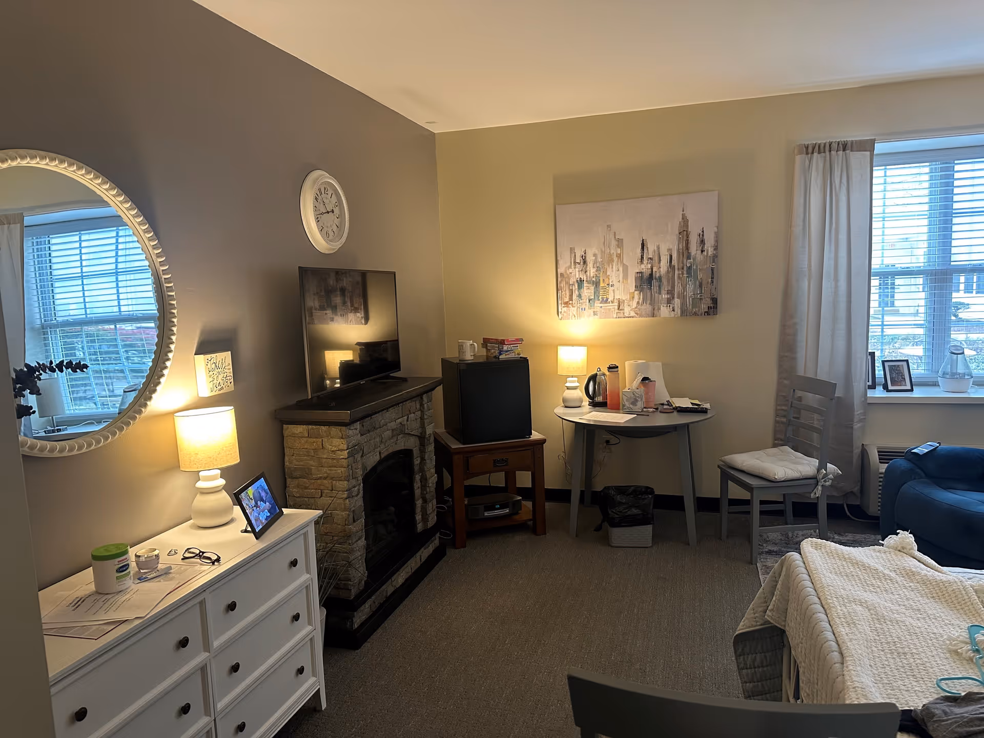 A cozy senior living room featuring a TV on a stone fireplace mantel, a round table with a lamp and various items, a white dresser with a lamp and framed photo, a large round mirror on the wall, a window with blinds and curtains, a blue armchair, and a bed partially visible in the foreground.