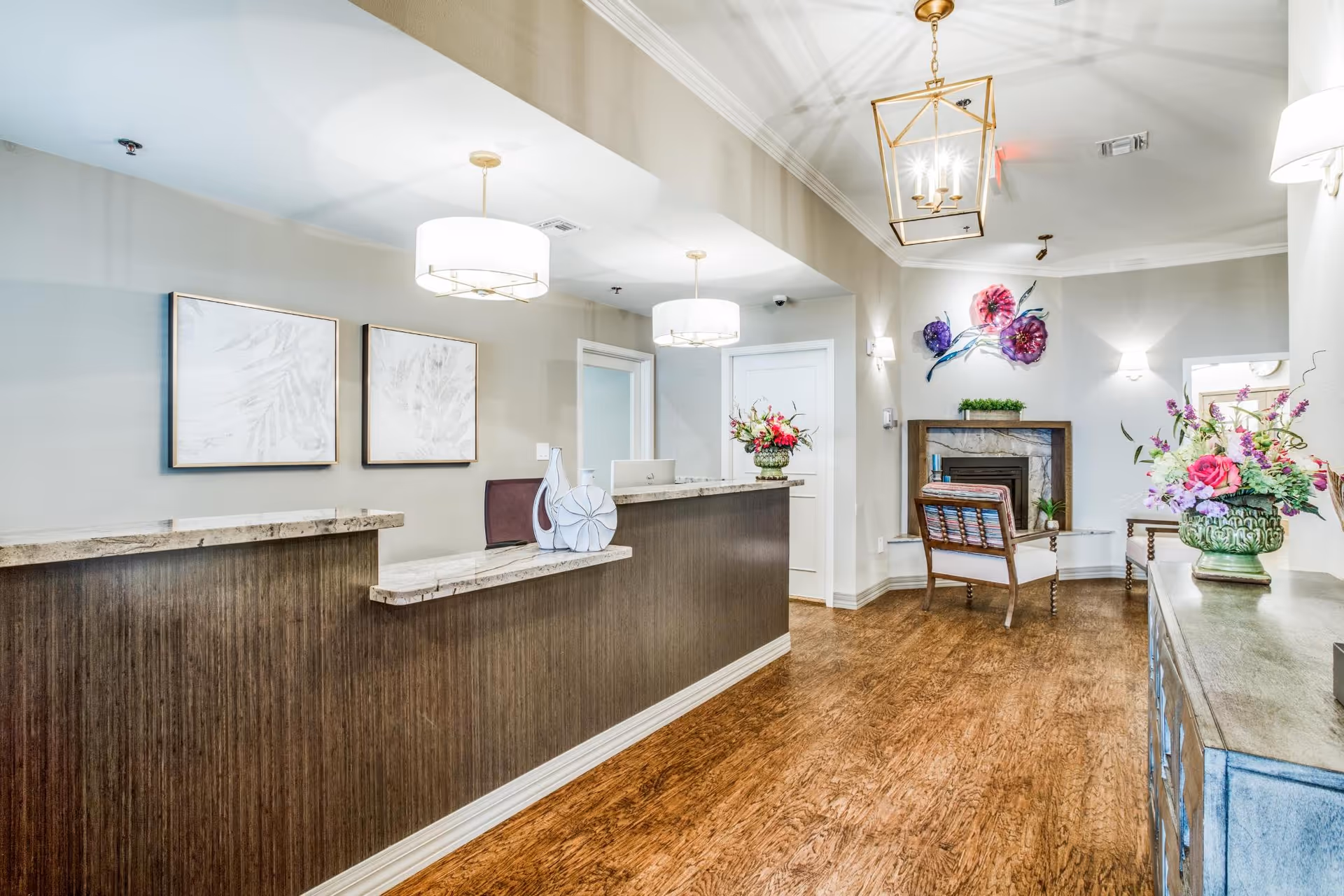 Reception area of Iris Memory Care of Nichols Hills featuring a wooden front desk with marble countertop, two modern ceiling lights, decorative vases with flowers, two framed artworks on the wall, a seating area with a chair near a fireplace, and floral wall decorations.