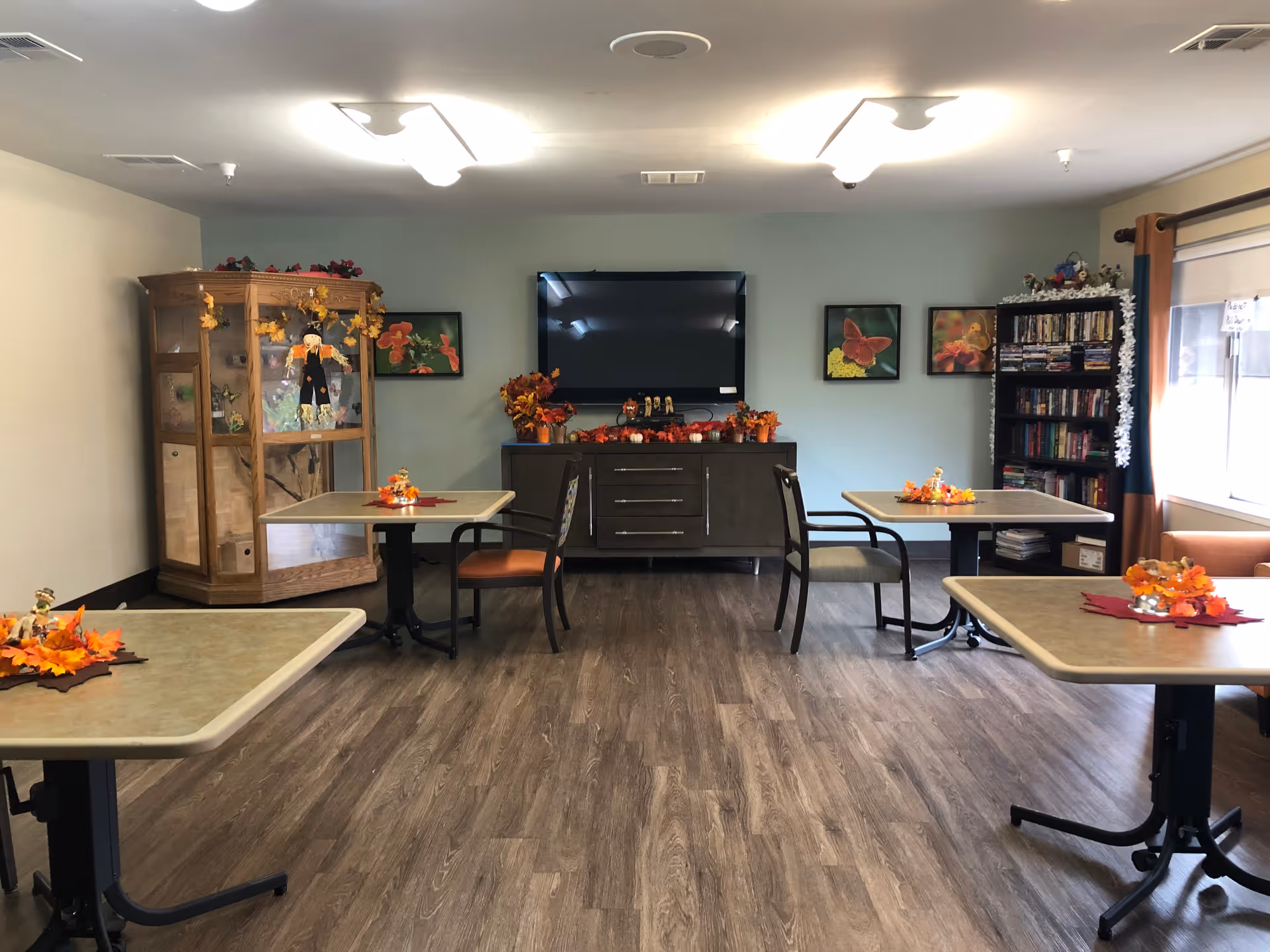 A common area in a senior living facility with four tables, each decorated with autumn-themed centerpieces. There are chairs around the tables, a wooden display cabinet with decorations on the left, a flat-screen TV mounted on a light blue wall, and a dark cabinet beneath the TV adorned with fall decorations. On the right side, there is a bookshelf filled with books and DVDs next to a window with curtains.