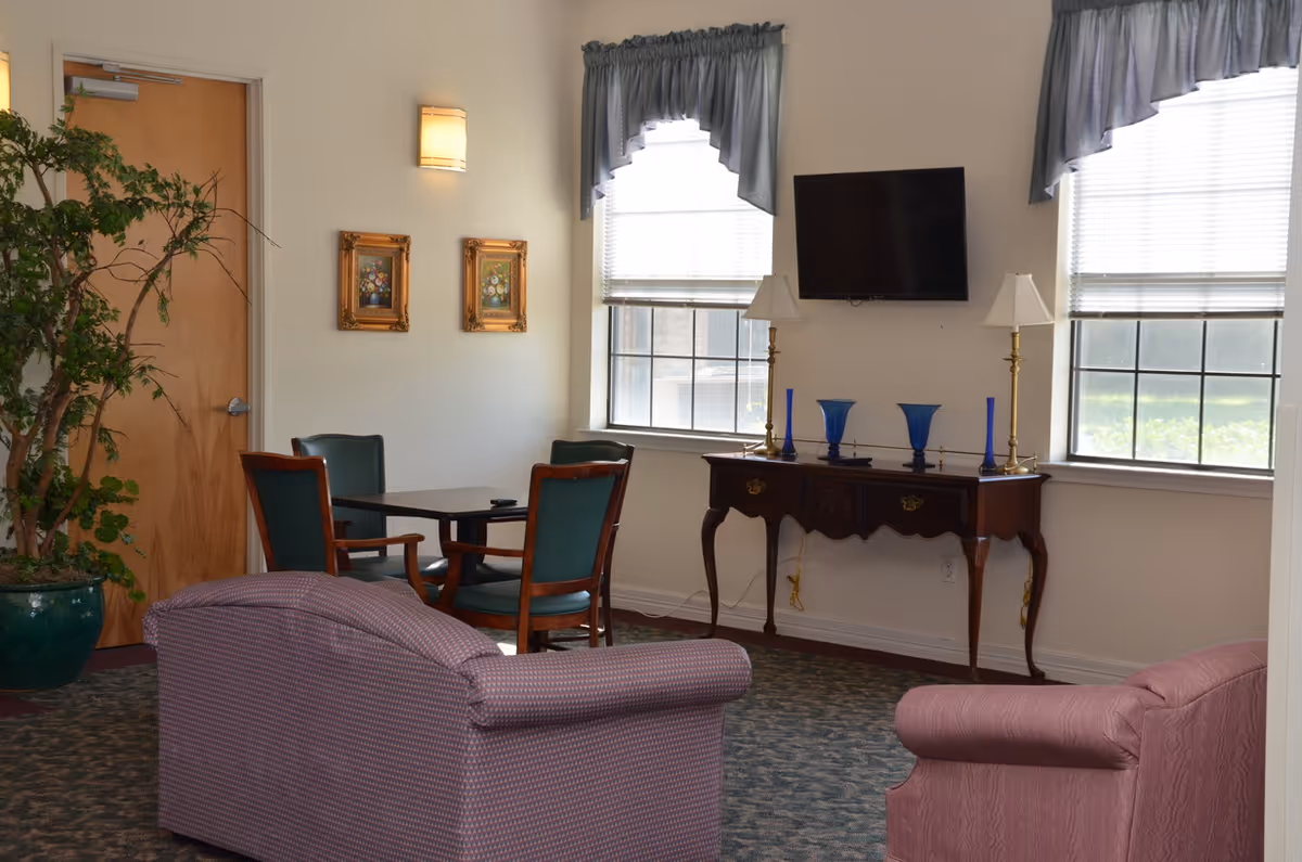 A cozy living room area with two upholstered armchairs and a small table with four chairs. There is a wooden console table against the wall with decorative blue vases and two lamps. A flat-screen TV is mounted on the wall between two windows with blue valances. A potted plant is near a wooden door, and two framed floral paintings hang on the wall.
