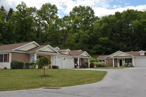 Single-story residential buildings with garages in a senior living community, surrounded by green trees and a well-maintained lawn under a partly cloudy sky.