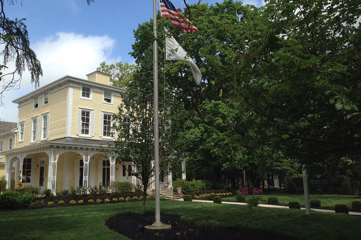 A large yellow three-story building with white trim and decorative porch columns, surrounded by green trees and a well-maintained lawn. Two flags, including an American flag, are flying on a flagpole in front of the building. A sign near the entrance reads 'Brandywine Senior Living at Haddonfield 132'.