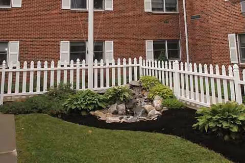 Brick senior living building with white picket fence, lawn, and a small rock waterfall feature.