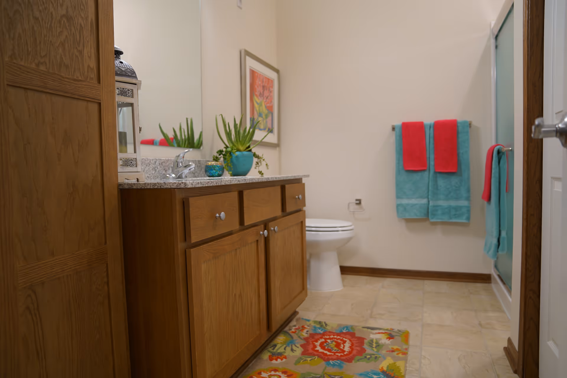 A bathroom with a wooden vanity cabinet topped with a granite countertop and a sink. There is a large mirror above the vanity, a toilet in the background, and colorful towels hanging on the wall. A decorative rug with a floral pattern is on the tiled floor, and a framed picture hangs on the wall.