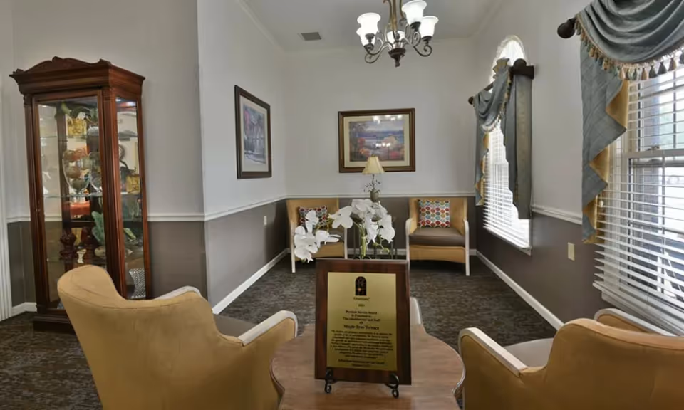 Small sitting room in an assisted living facility featuring upholstered chairs around a table, a glass display cabinet, framed wall art, and windows with draped curtains.