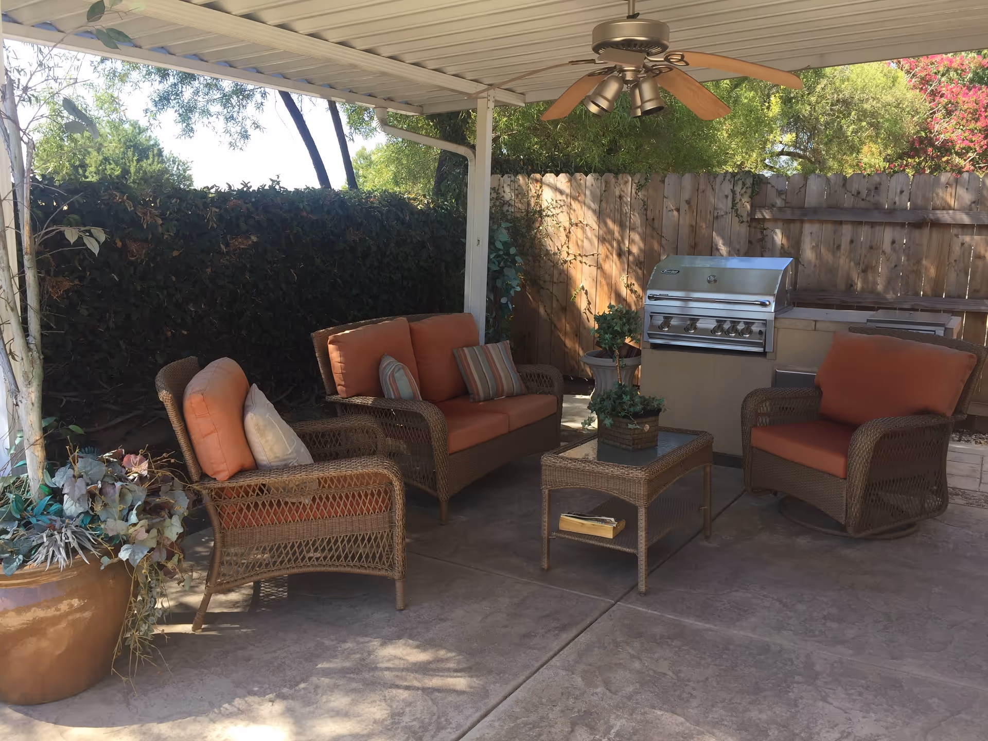 Outdoor covered patio area with wicker furniture including a loveseat, two armchairs with orange cushions, and a glass-top coffee table with a small plant. There is a ceiling fan above and a stainless steel barbecue grill against a wooden fence. Potted plants and greenery surround the space.