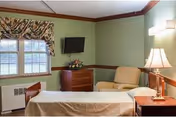 A single bedroom in a senior living facility with a bed covered in a beige blanket, a beige armchair, a wooden dresser with a flower arrangement, a wall-mounted flat-screen TV, a window with floral curtains, and a bedside table with a lamp.