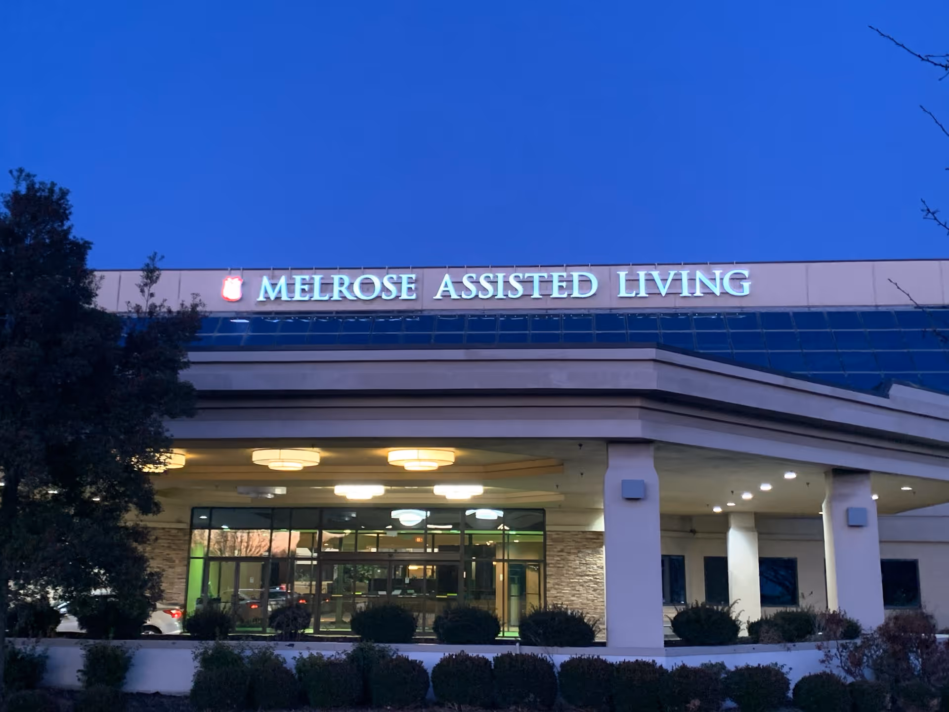 Exterior front view of Melrose Assisted Living facility at dusk with illuminated signage and entrance area surrounded by bushes and trees.
