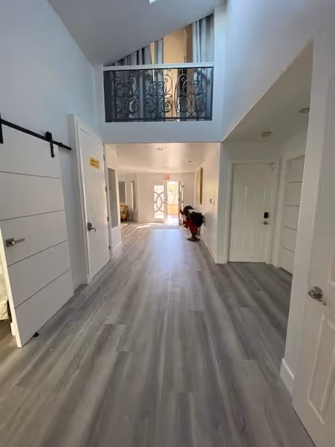 A bright hallway with light gray wood flooring and white walls. There are multiple white doors on both sides, a decorative black metal railing on the upper level, and a glass door at the far end letting in natural light. A small decorative figure is placed near the far wall.