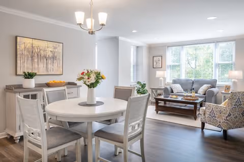 Open-plan senior living area with a round dining table in the foreground and a bright living room with sofa and armchair by large windows.