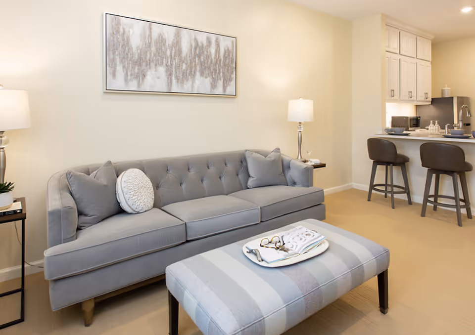Neutral-toned living room with a gray tufted sofa, striped ottoman with a tray, table lamps, and an open kitchen counter with bar stools.