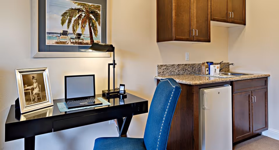 Studio kitchenette and workspace with a granite countertop, mini-fridge and sink on the right and a black desk with a laptop, framed photo and blue chair on the left.
