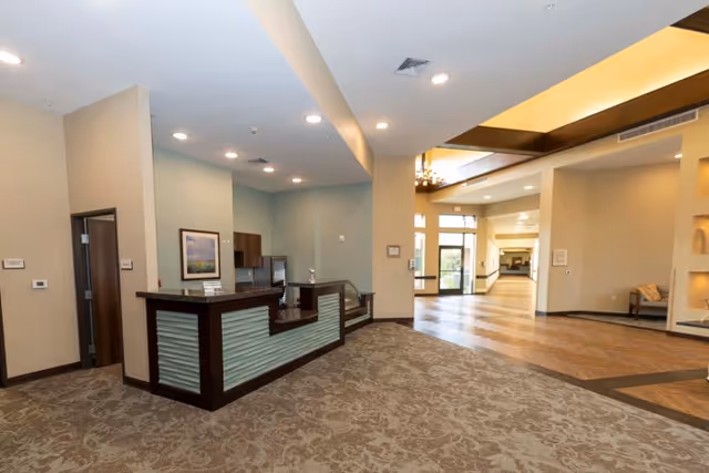Interior view of a senior living facility lobby area with a reception desk on the left, beige walls, patterned carpet, and a hallway leading to glass doors and additional seating areas.