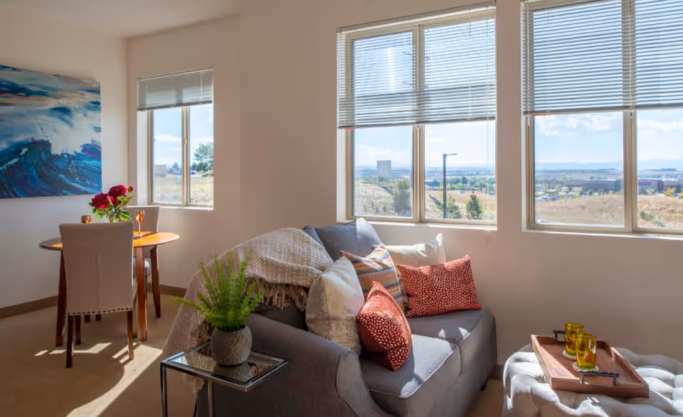A bright living room area with a gray couch adorned with multiple colorful pillows and a throw blanket. Next to the couch is a small glass side table with a potted fern. In the background, there is a small round wooden dining table with two beige upholstered chairs and a vase with red flowers. Large windows with blinds allow natural light to fill the room and offer a view of an outdoor landscape with hills and buildings. A large abstract painting with blue tones hangs on the wall near the dining area.