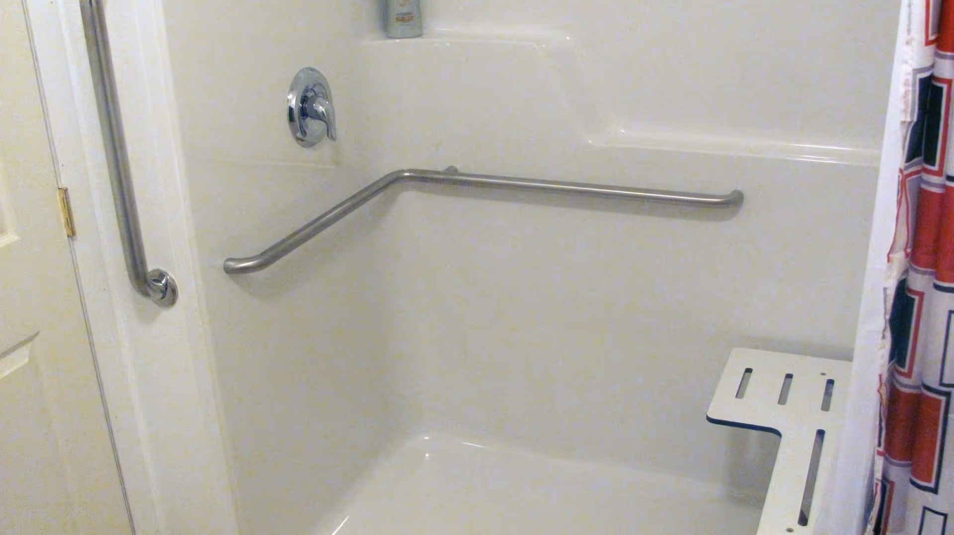 A white shower area with a built-in seat and two stainless steel grab bars, one vertical near the door and one horizontal along the back wall. A shower curtain with a red, white, and blue geometric pattern is partially visible on the right side.