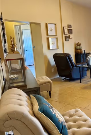 Interior view of a living room area with a beige tufted sofa adorned with decorative pillows in the foreground. A console table with a large lantern and decorative items is positioned against the wall. In the background, there is a dark recliner chair next to a small round table, and wall decorations including framed art and plaques. The floor is tiled and the walls are painted a light beige color.