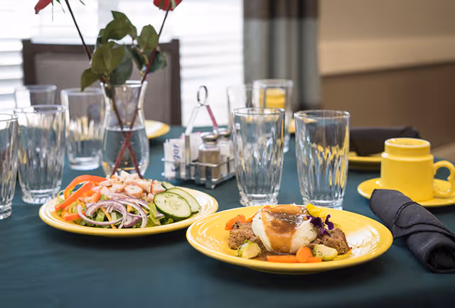 A dining table set with two yellow plates of food, one with a salad including cucumbers, tomatoes, and onions, and the other with a main dish featuring meatloaf, mashed potatoes with gravy, and vegetables. The table also has several empty drinking glasses, a yellow cup and saucer, black cloth napkins, and a vase with red flowers in the background.