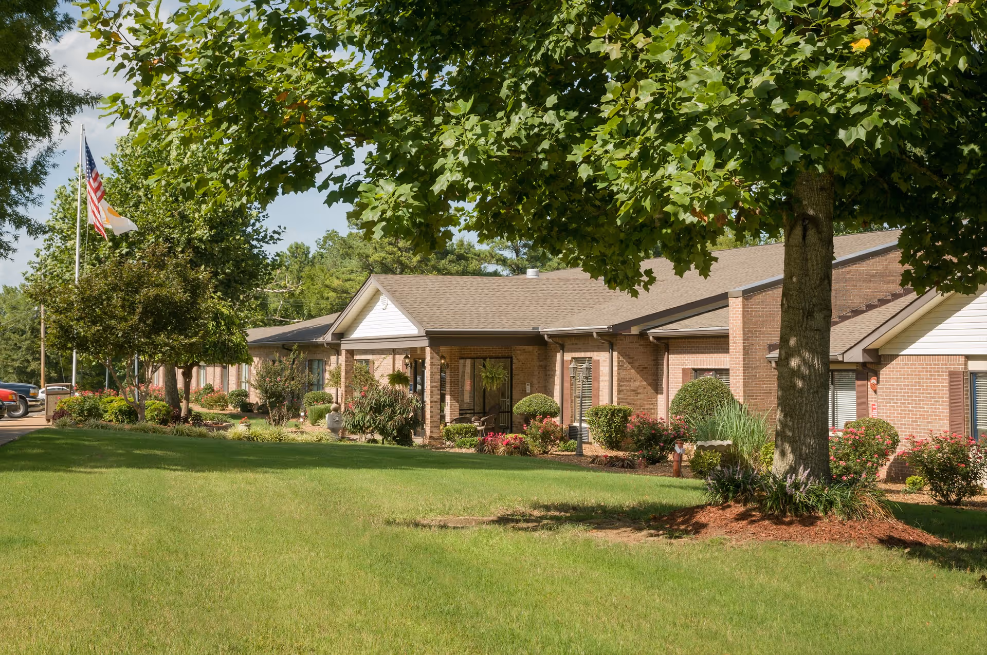 Single-story brick senior living facility building with a manicured lawn, trees, and flags in front.