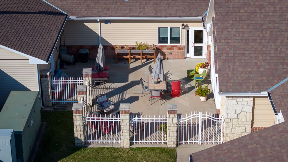 Fenced outdoor courtyard patio between buildings with tables, chairs, umbrellas and potted plants.