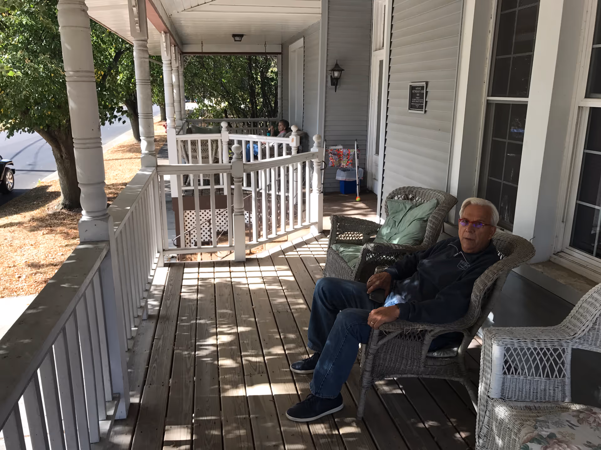 An elderly man sitting on a wicker chair on a wooden porch with white railings and columns. There are other chairs and a small table with a floral cushion on the porch. Trees and a parked car are visible in the background.