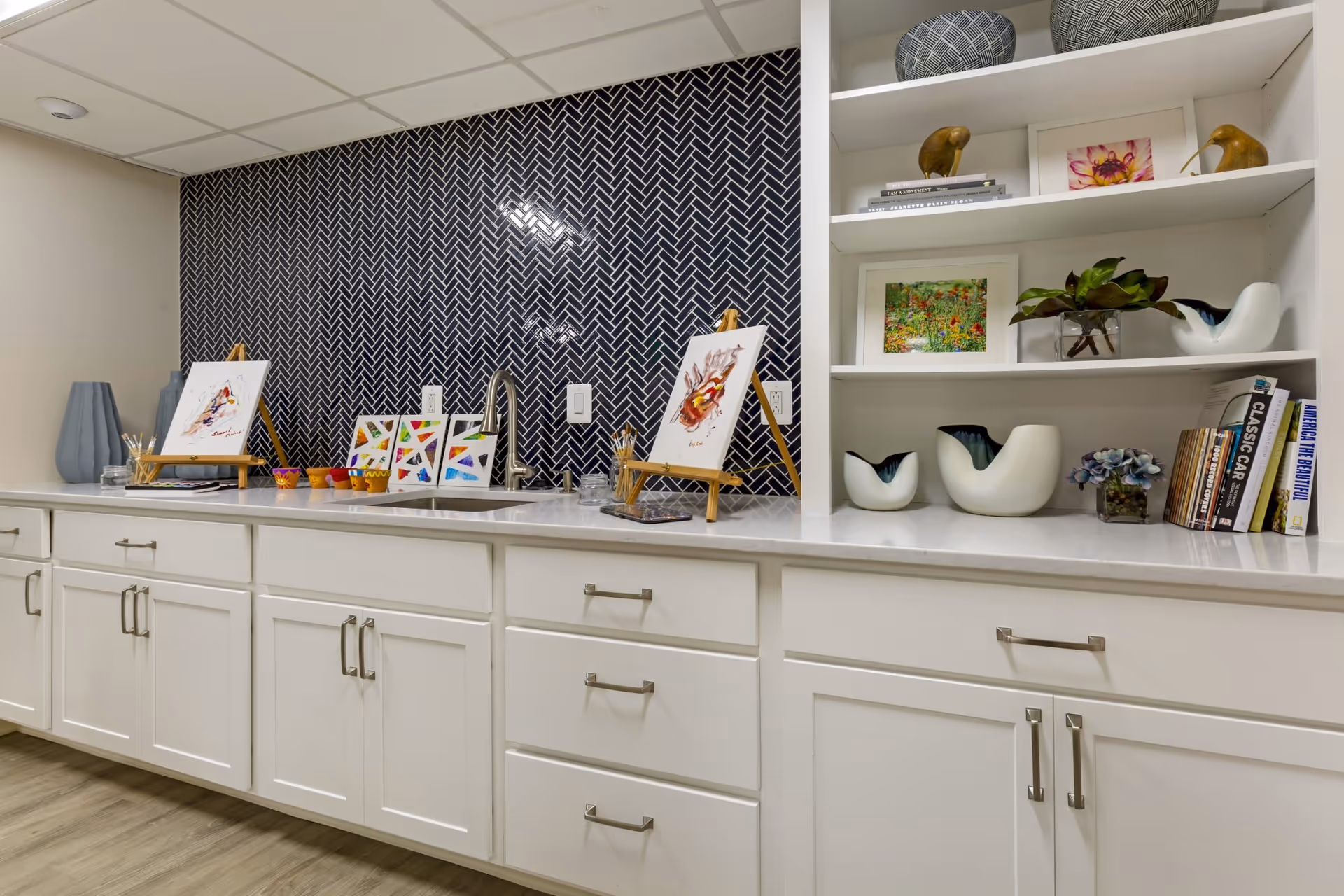 A countertop with white cabinets below and open shelves above, decorated with small paintings on easels, colorful paint pots, books, ceramic bird figurines, and a plant. The backsplash features a dark tile with a herringbone pattern, and there is a sink with a modern faucet in the center.