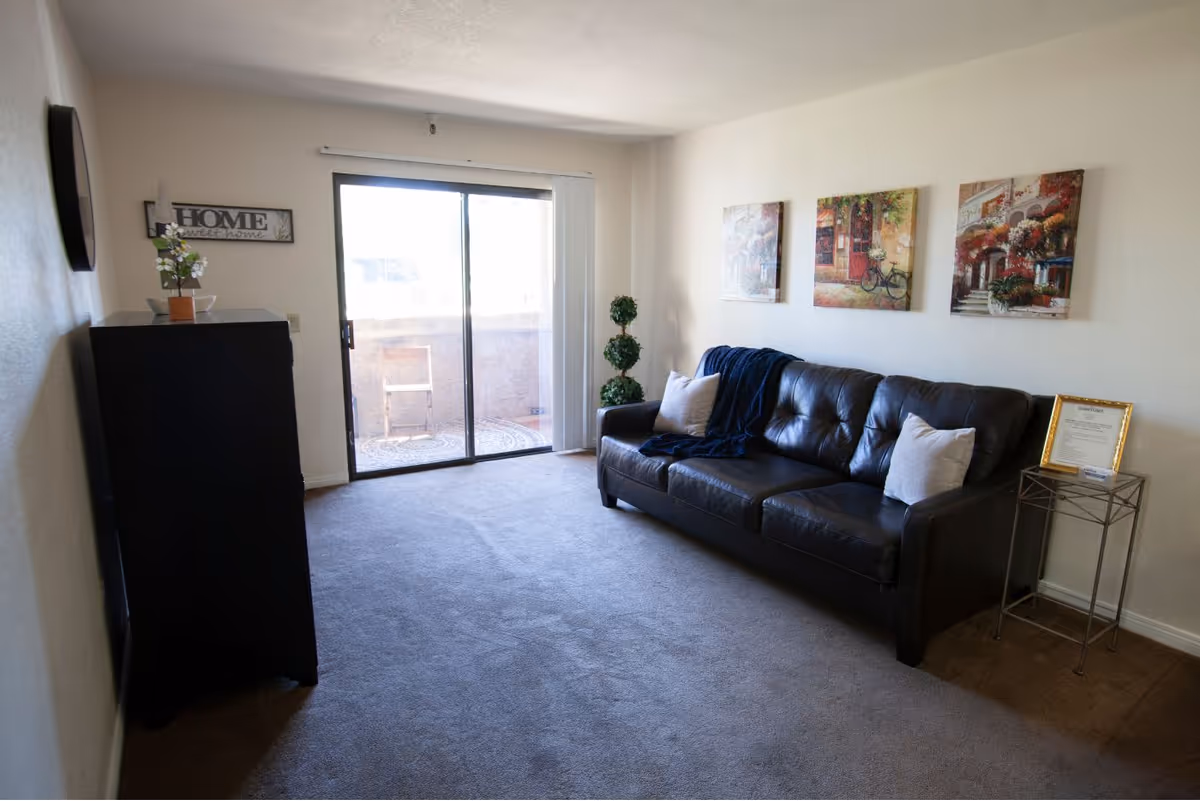 A living room with a black leather sofa adorned with two light-colored pillows and a dark blue throw blanket. Above the sofa are three colorful paintings depicting outdoor scenes. To the right of the sofa is a small metal side table with a framed document and a small box on it. On the left side of the room is a black cabinet with a small plant on top and a wall decoration that reads 'HOME sweet home'. The room has carpeted flooring and a sliding glass door leading to a small balcony with a chair outside.