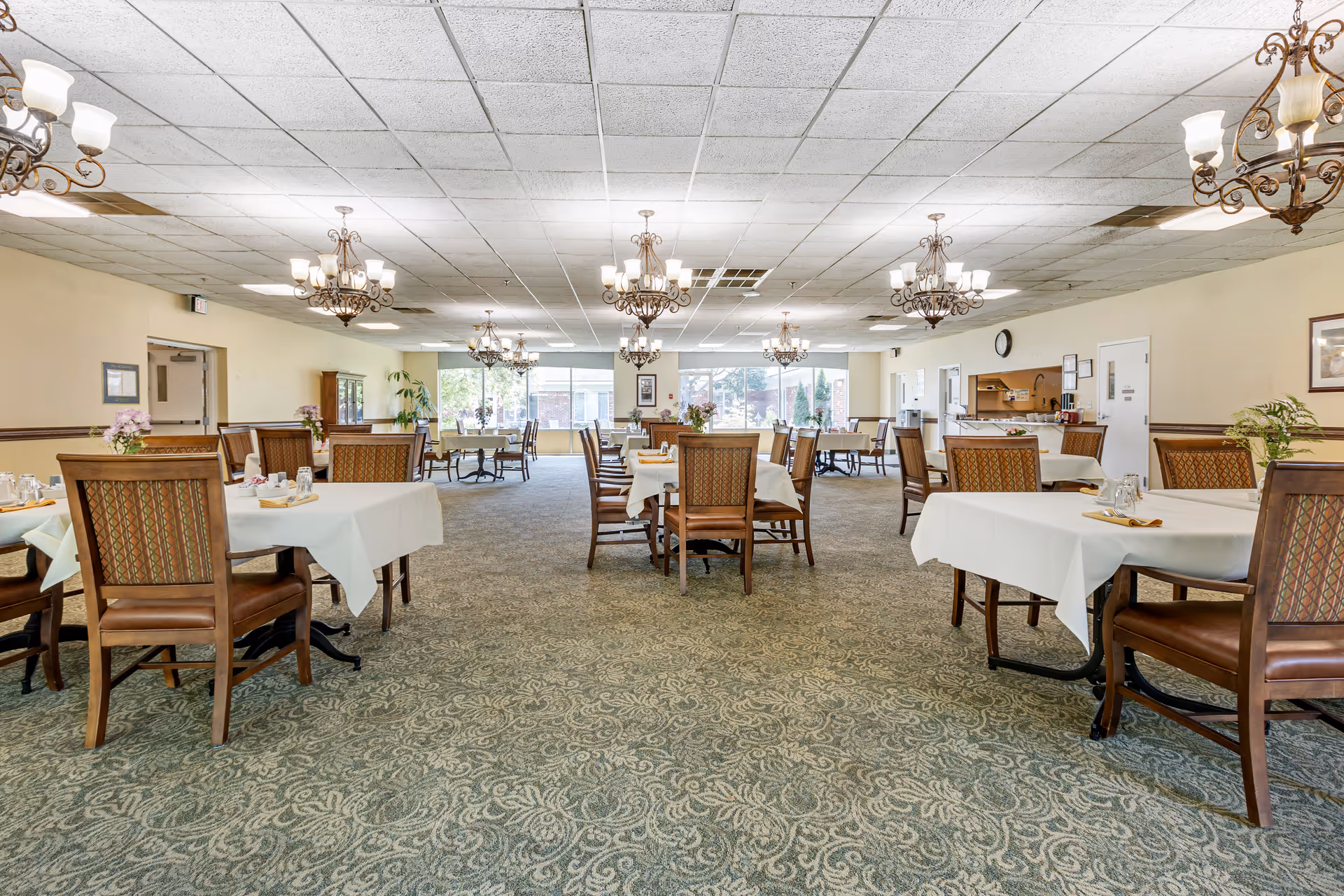 A spacious dining room with multiple tables covered in white tablecloths, each surrounded by wooden chairs with patterned upholstery. The room has a patterned carpet, several ornate chandeliers hanging from the ceiling, and large windows at the far end letting in natural light. There are flowers on the tables and a counter area on the right side of the room.