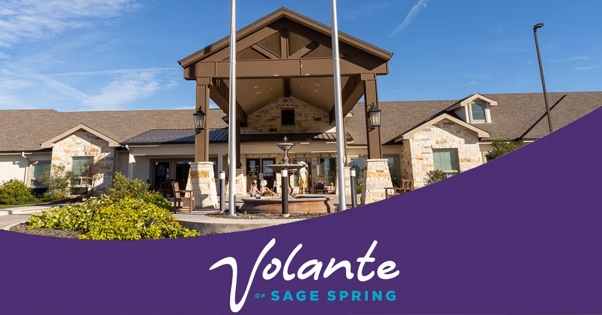 Front exterior view of Volante Senior Living of Sage Spring building with stone and wood facade, a covered entrance with pillars, a fountain, and landscaping under a clear blue sky.