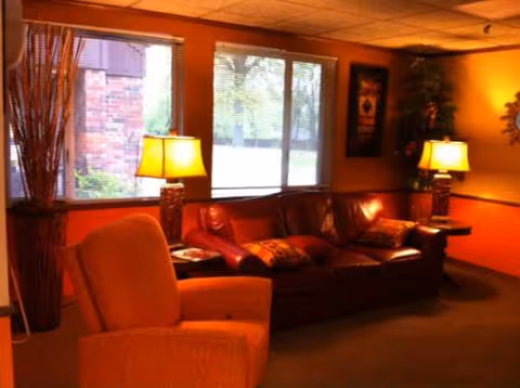 Warm senior living lounge with leather sofa, armchair, lamps, and large windows.