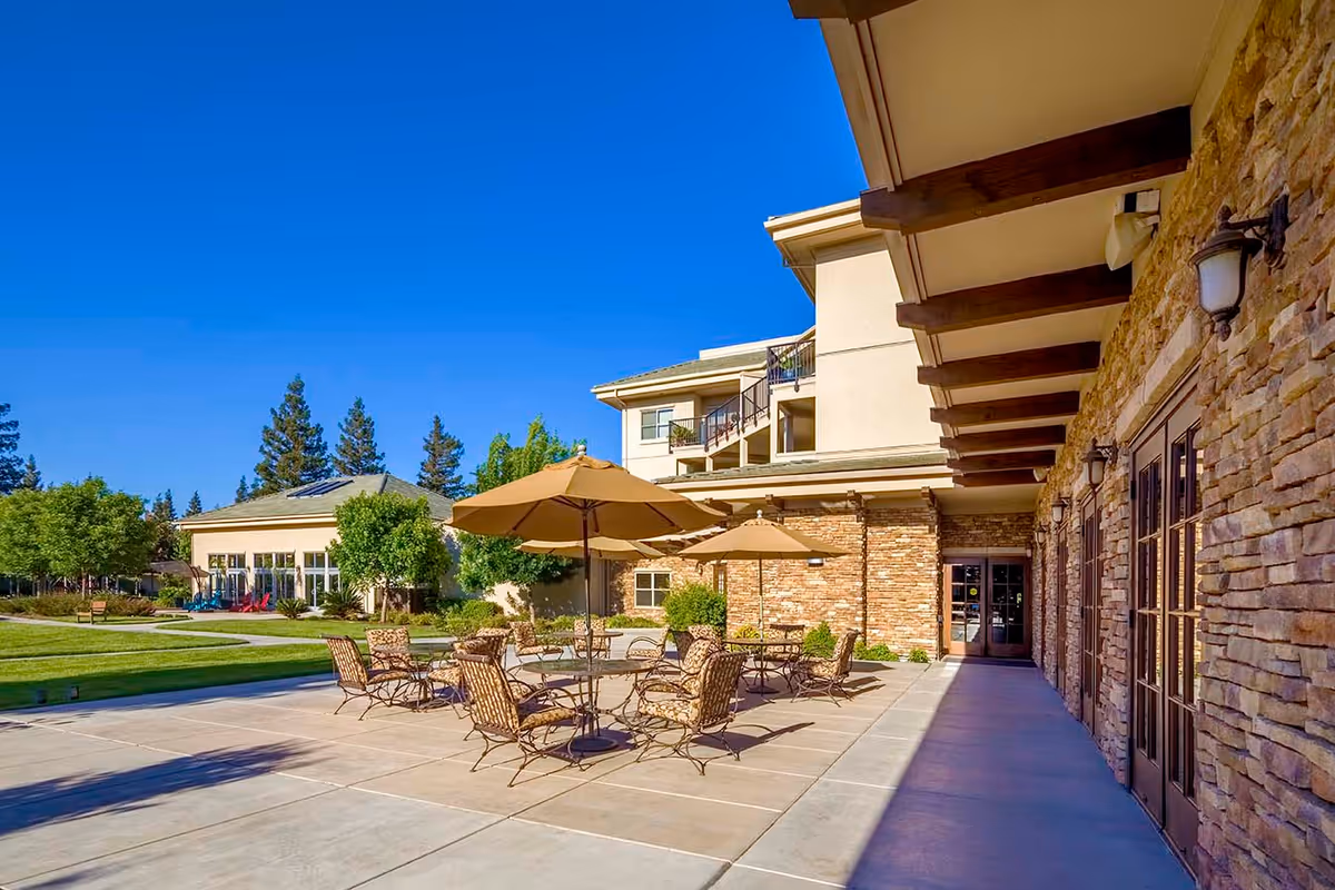 Sunny outdoor patio with tables, chairs and umbrellas beside a stone-clad senior living building and lawn.