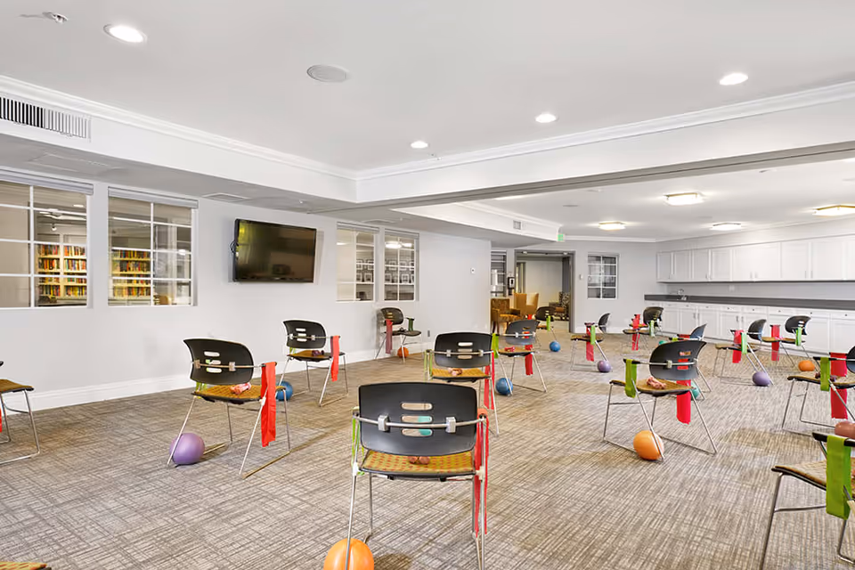 A spacious activity room with multiple chairs arranged in rows, each chair equipped with colorful resistance bands and small exercise balls on the floor nearby. The room has a carpeted floor, white walls, a wall-mounted TV, and windows looking into another room with bookshelves. There are white cabinets and countertops along one wall, and the ceiling has recessed lighting.