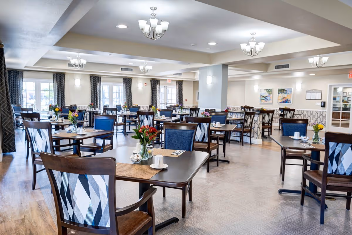 A bright and spacious dining room with multiple wooden tables and chairs arranged neatly. Each table has a small vase with colorful flowers and place settings including cups and napkins. The room features large windows with patterned curtains, light-colored walls, and ceiling lights with chandeliers providing illumination.