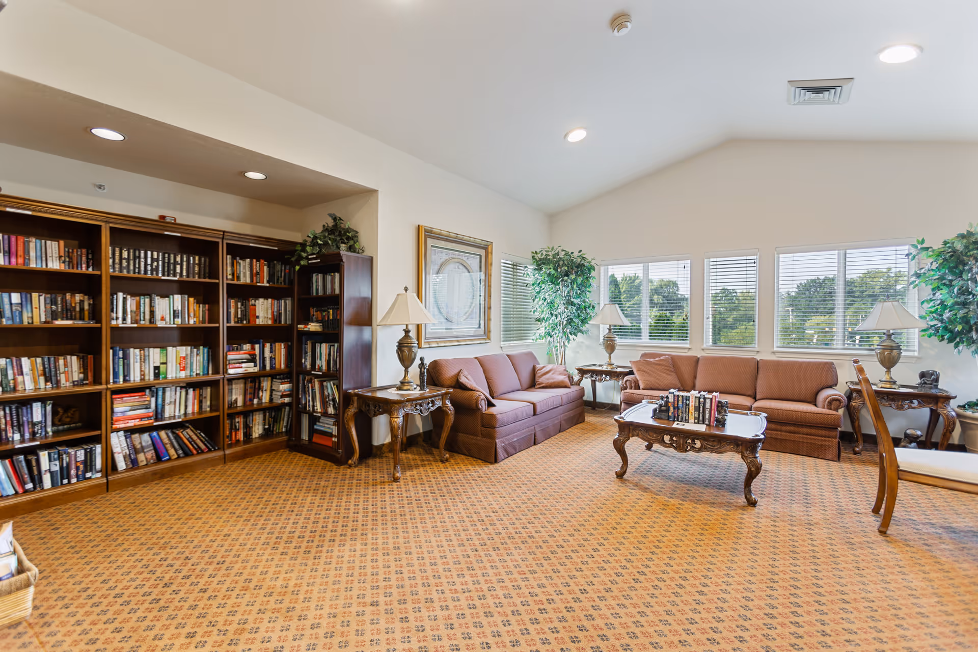 A bright and spacious living room area with two brown sofas, a wooden coffee table with books on it, two side tables with lamps, large windows with blinds letting in natural light, and a large bookshelf filled with books along one wall. The room has a patterned carpet and decorative plants near the windows.