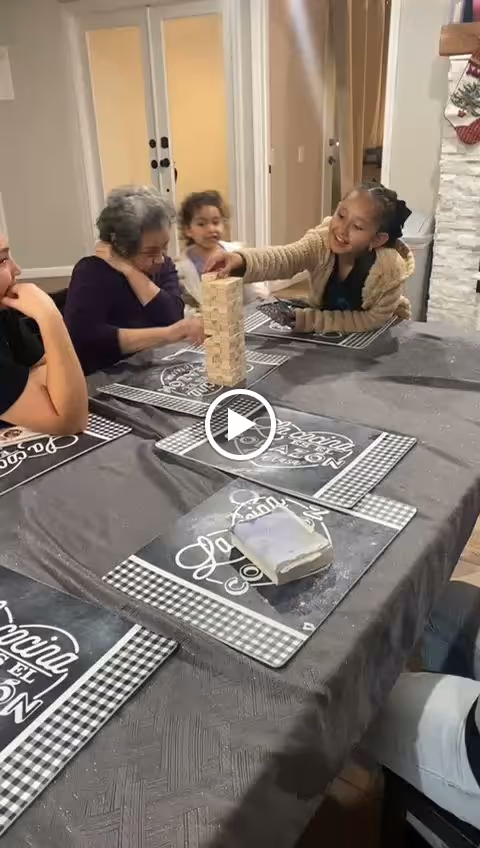 A group of people, including an elderly woman and children, sitting around a dining table playing a game of Jenga. The table is covered with a gray tablecloth and has black and white placemats with text on them. The setting appears to be a cozy indoor dining area with a fireplace and double doors in the background.