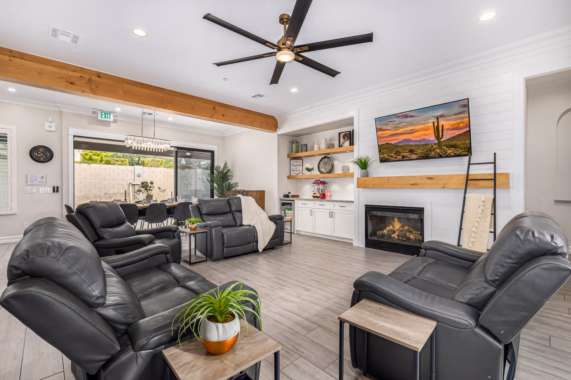 Bright common lounge with multiple black leather recliners arranged around a fireplace, wall-mounted TV, built-in shelving and a dining area visible through sliding glass doors.