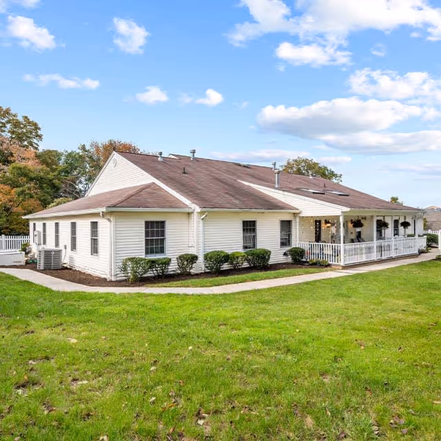 Single-story building with white siding and a brown roof, surrounded by green grass and a few small bushes. The building has multiple windows and a covered porch area with hanging plants. The sky is blue with scattered clouds.