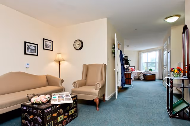 Interior view of a senior living facility room with a beige sofa and armchair, a floor lamp, and framed pictures on the wall. A trunk-style coffee table with decorative items is in front of the sofa. The room extends into a bedroom area with a bed near a large window letting in natural light. A small table with a mirror and flowers is on the right side of the image.