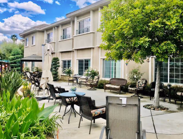 Outdoor patio area at Villas at San Bernardino with multiple chairs, tables, umbrellas, and greenery including a tree and plants along the walkway beside a beige two-story building under a partly cloudy sky.