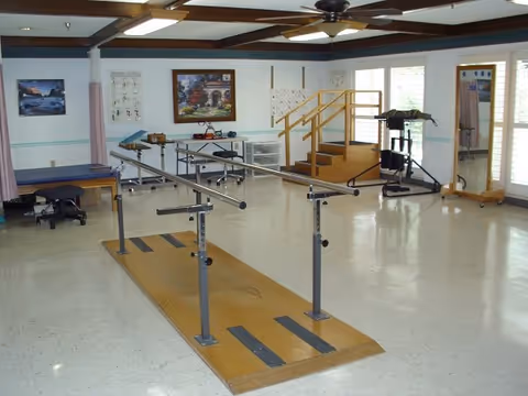 A spacious rehabilitation room with parallel bars for walking exercises on a wooden platform, a small set of stairs with handrails, a treatment table, exercise equipment, and large windows letting in natural light.