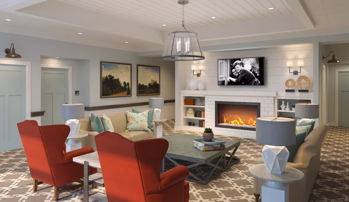 A cozy living room in Church Hill Village featuring two red armchairs, a beige sofa with floral and solid teal pillows, a large coffee table with books and a small plant, a modern fireplace with a fire burning, built-in shelves with decorative items and books, a wall-mounted TV displaying a black and white image of two people hugging, two landscape paintings on the wall, and soft lighting from lamps and ceiling lights.