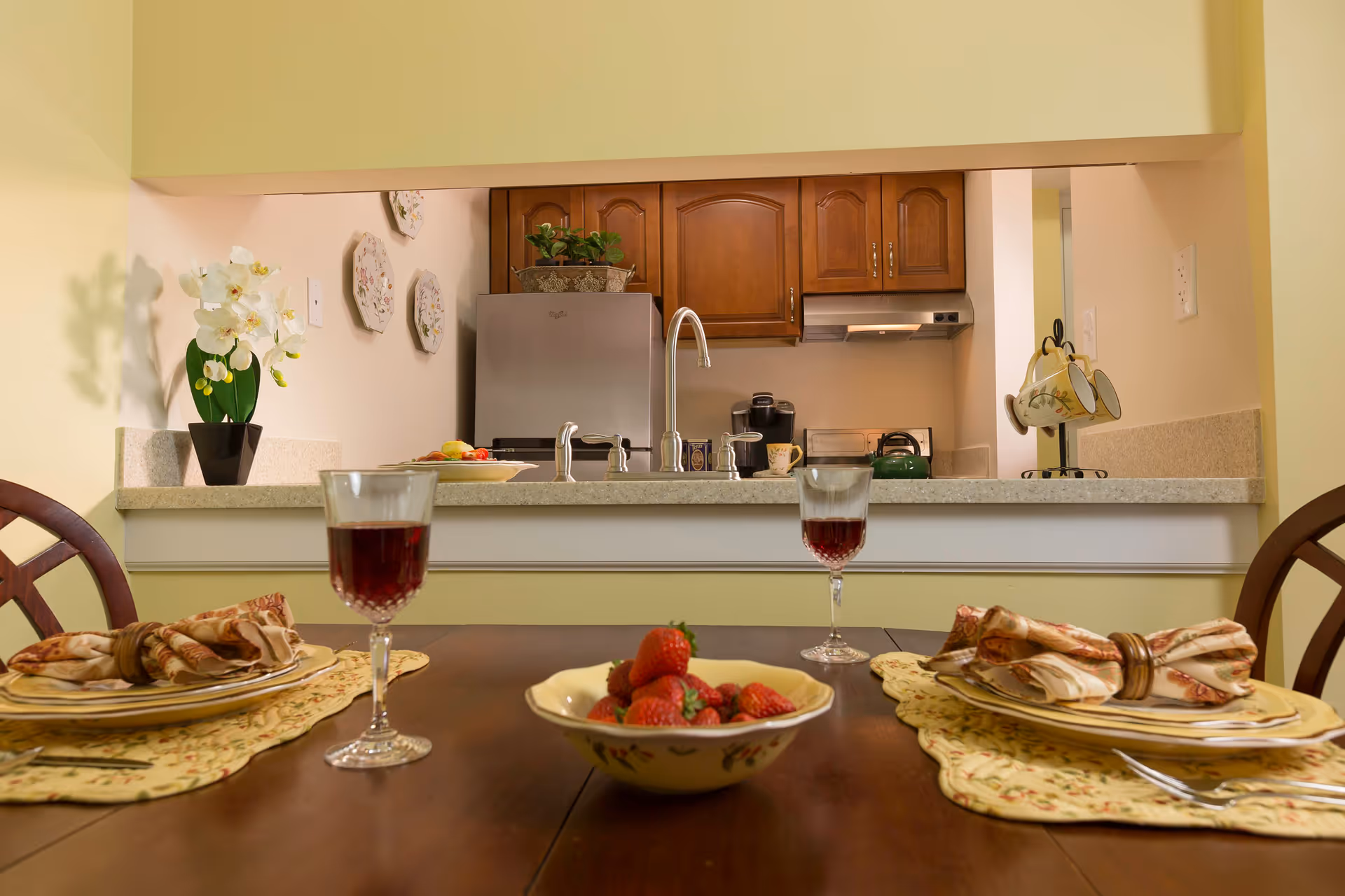 A dining table set for two with plates, napkins, and glasses of red wine. In the background, there is a kitchen with wooden cabinets, a stainless steel refrigerator, a sink, and a countertop with a bowl of fruit and a coffee maker. The walls are decorated with floral plates and a potted orchid plant.