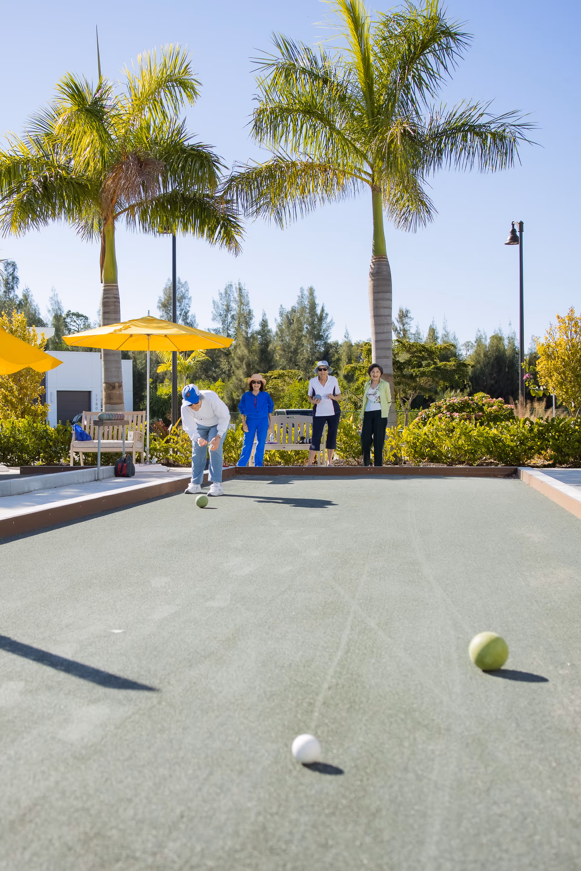 Four seniors playing bocce ball on an outdoor court surrounded by palm trees and greenery under a clear blue sky. Two yellow umbrellas and benches are visible in the background.