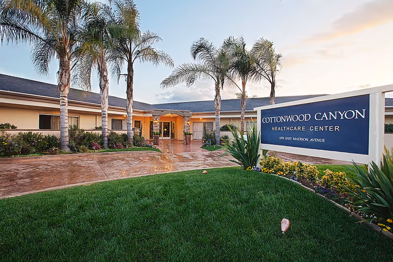 Exterior view of Cottonwood Canyon Healthcare Center showing the entrance with a pathway, palm trees, and a large sign displaying the facility's name and address. The sky is partly cloudy with soft lighting suggesting early morning or late afternoon.