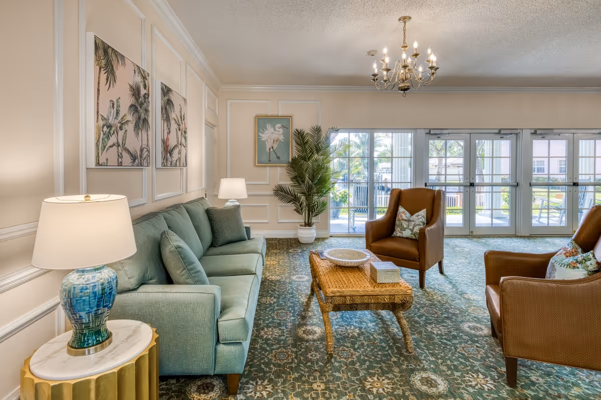 A cozy living room with a light green sofa, two brown armchairs with floral pillows, a wicker coffee table with decorative items, a blue and white table lamp on a round side table, tropical-themed wall art, a potted plant, and large glass doors leading to an outdoor area.