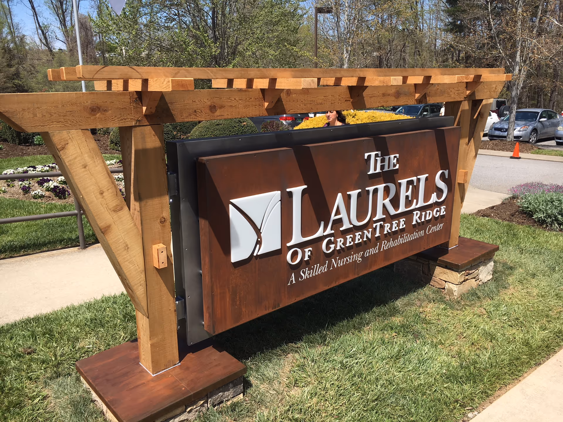 Outdoor wooden sign for The Laurels of GreenTree Ridge, a skilled nursing and rehabilitation center, set on a grassy area with a sidewalk and parked cars in the background.